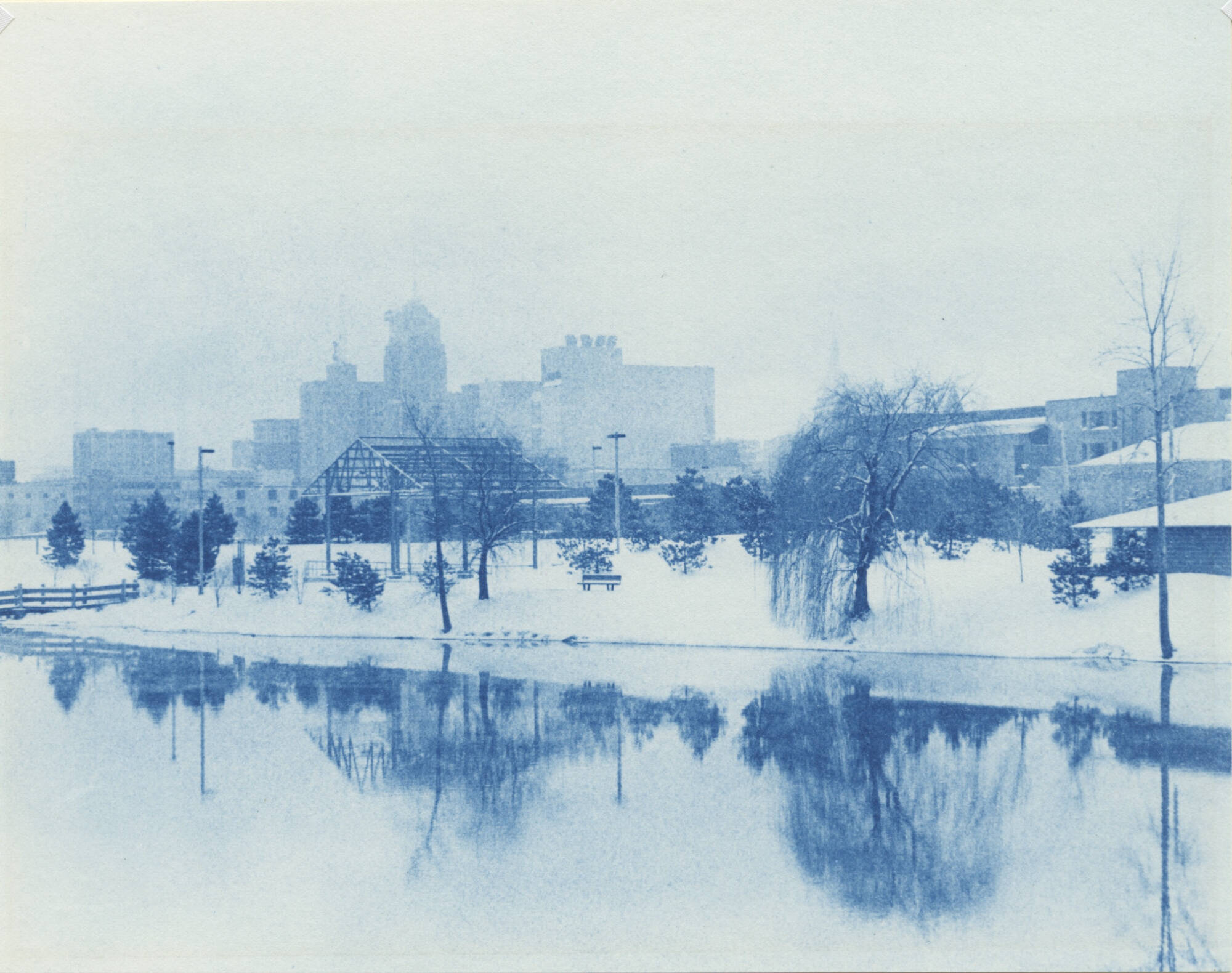Cyanotype image of buildings and trees reflected in a still body of water.